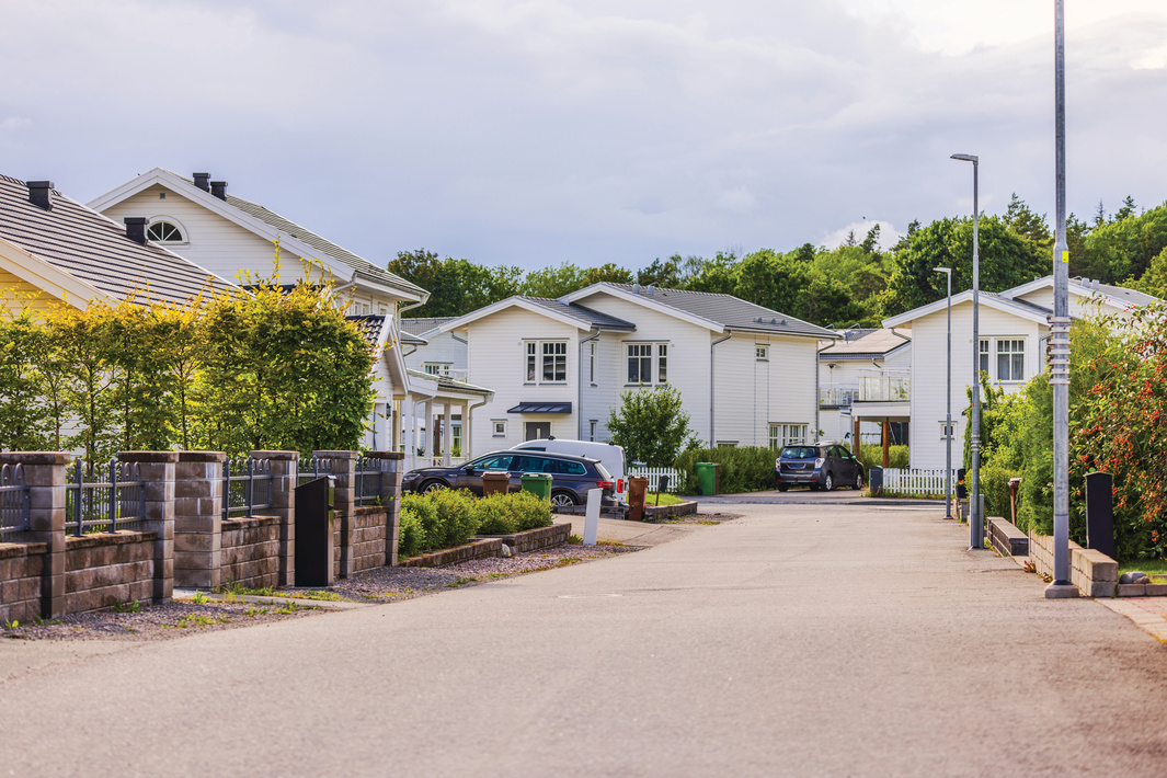 View of residential street with white houses, greenery, and parked cars in suburban area on sunny day. Sweden, Europe.